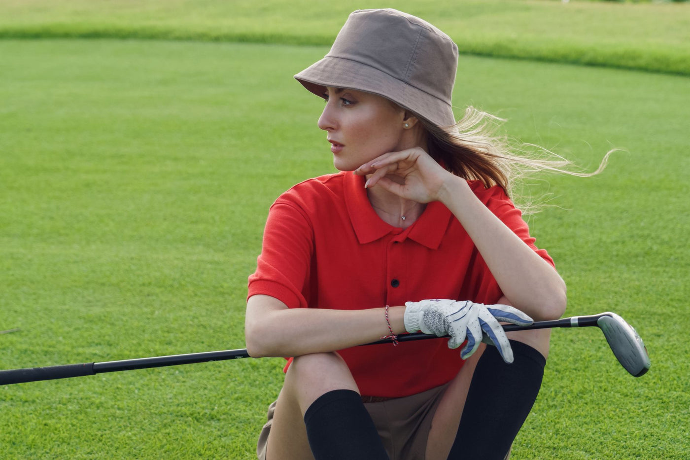 Fashionable woman in a red polo and hat sitting with a golf club on a lush green course.